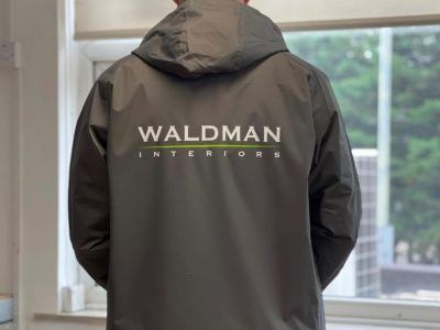 Baksidan av en mörkgrå profiljacka med trycket Waldman Interiors.
